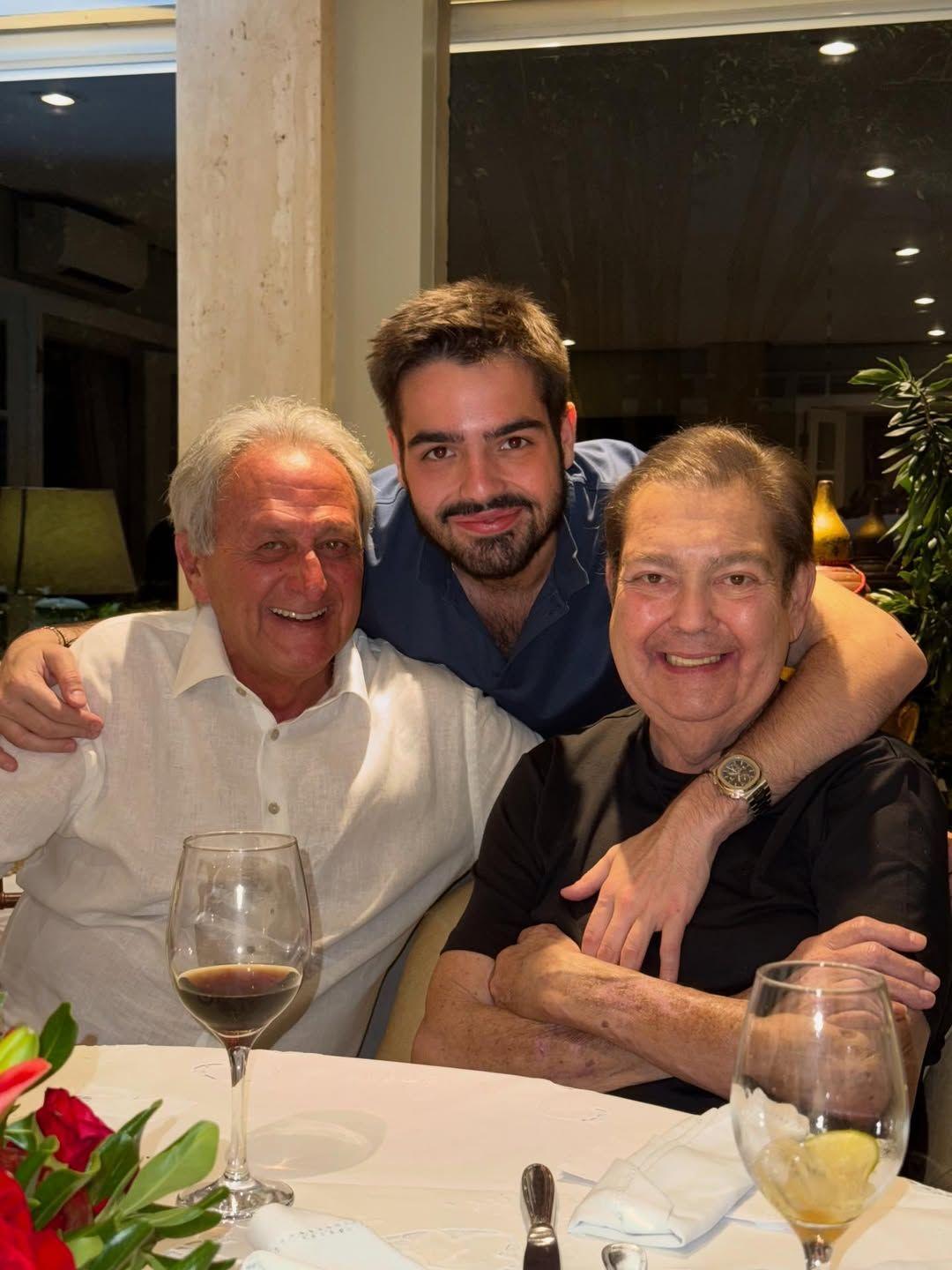 Retrato de família mostrando Fausto Silva, o filho e outro homem felizes à mesa, com taças de vinho.