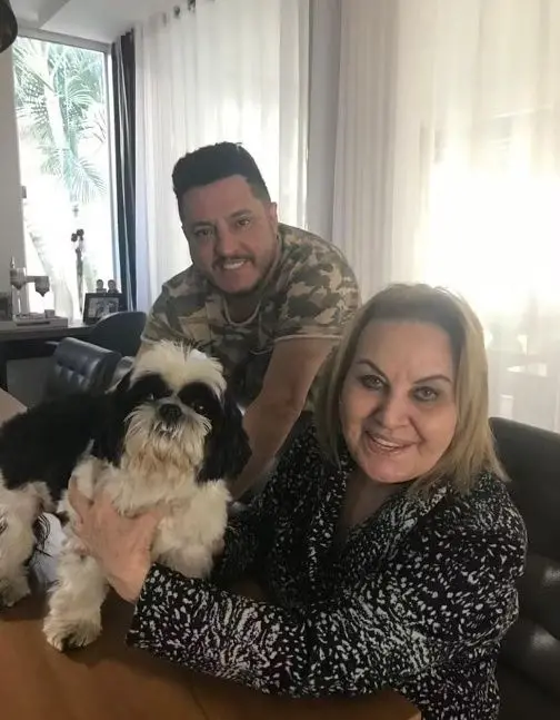 Bruno e a mãe, dona Anita, sorrindo com um pequeno cachorro Shih Tzu preto e branco em ambiente doméstico.