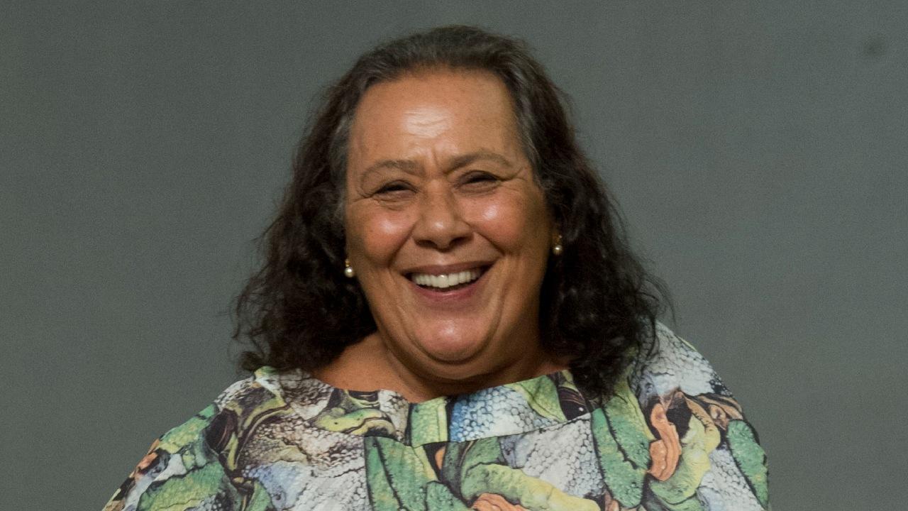 Teuda Bara em foto feita em estúdio. Ela está de vestido longo. Tem cabelos longos e grisalhos. Na foto, ela sorrir.