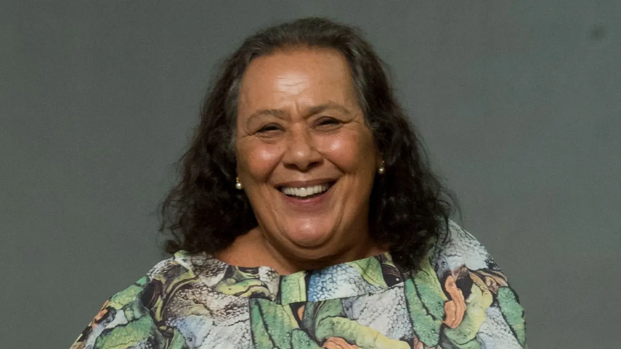 Teuda Bara em foto feita em estúdio. Ela está de vestido longo. Tem cabelos longos e grisalhos. Na foto, ela sorrir.