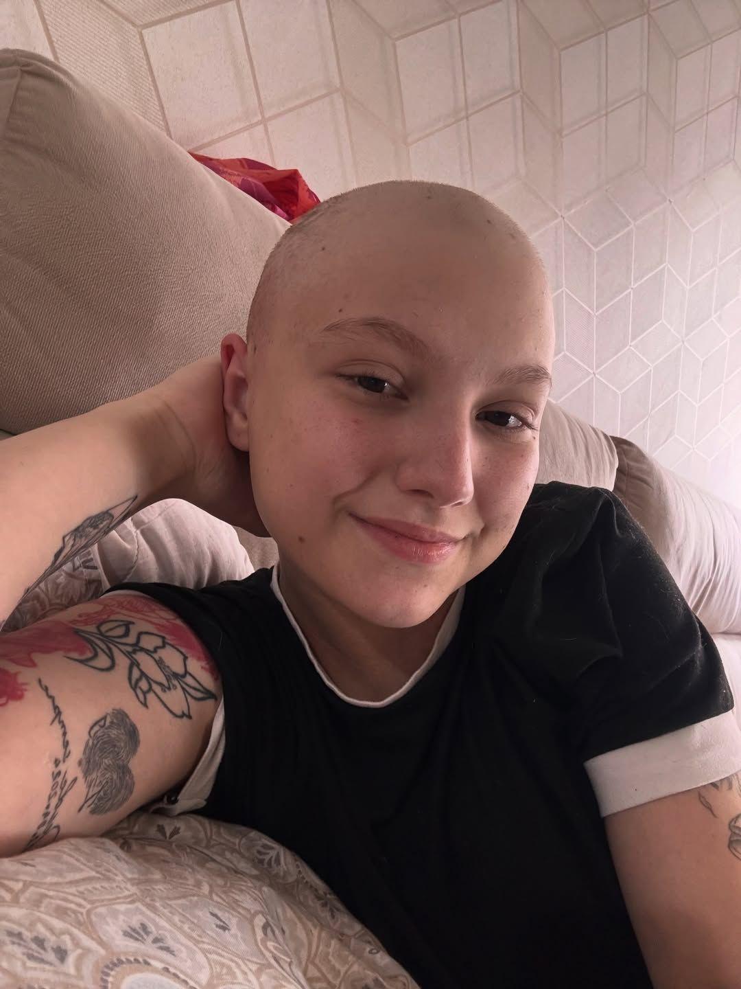 Isabel Veloso, sorridente com cabelo raspado e tatuagens no braço, apoiada em um sofá ou almofada.