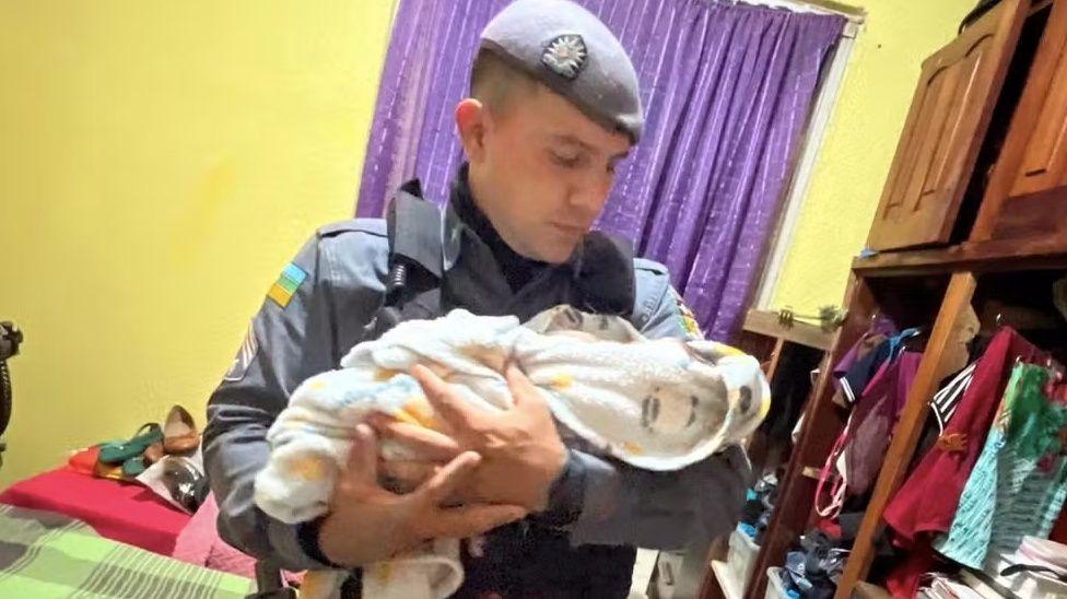 Um policial uniformizado, usando uma boina cinza, segura um bebê delicadamente nos braços, olhando para ele com ternura em um ambiente doméstico.