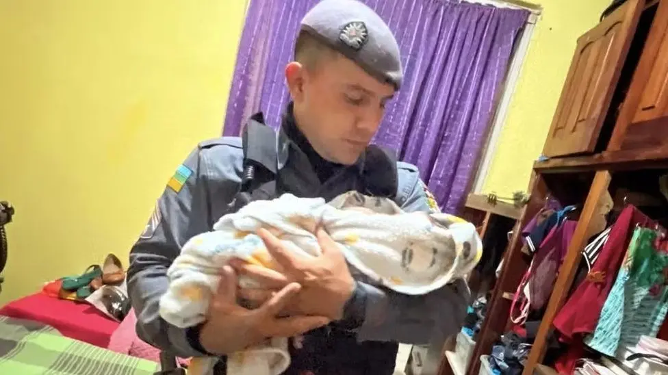 Um policial uniformizado, usando uma boina cinza, segura um bebê delicadamente nos braços, olhando para ele com ternura em um ambiente doméstico.
