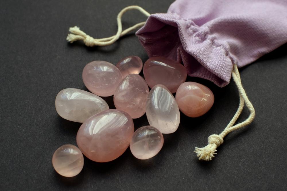Conjunto de pedras de quartzo rosa em uma superfície escura ao lado de uma pequena maleta de pano roxa com fechamento de cordão.