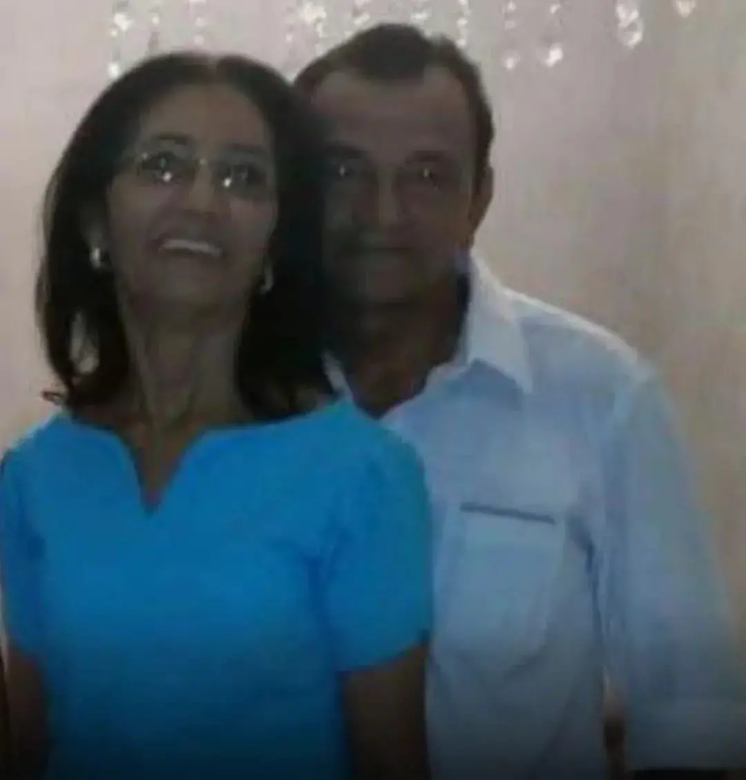 Imagem de casal sorridente, mulher de cabelo escuro e óculos, vestida com blusa azul, ao lado de homem de cabelo castanho e camisa branca, em ambiente de festa com fundo decorado.