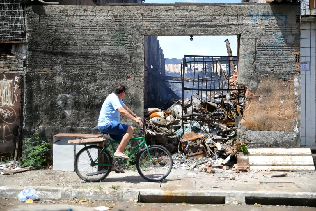 Homem de blusa azul e shorts em uma bicicleta, pedalando da esquerda para a direita e olhando para um muro atrás, com sucata queimada.