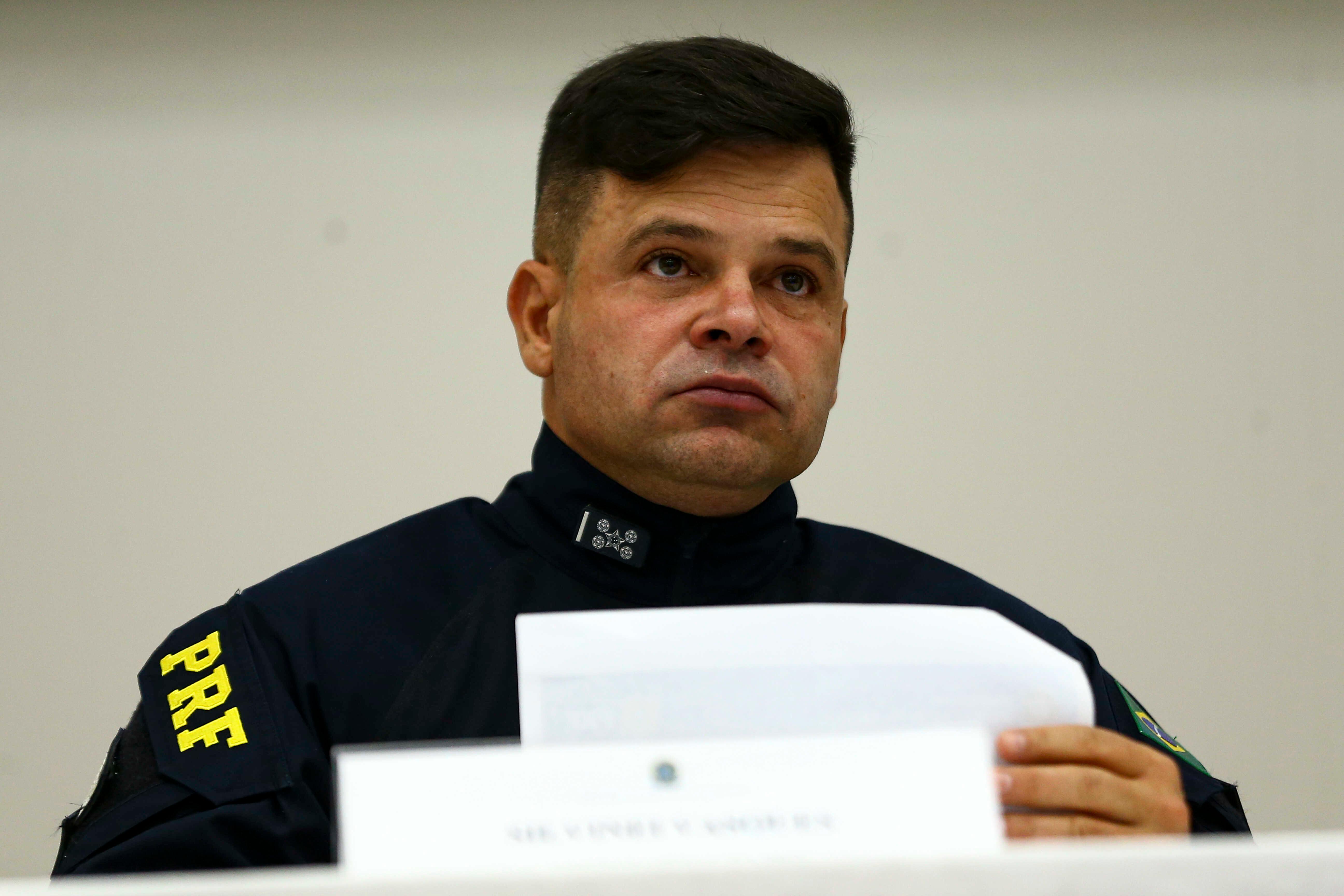 Homem usando uniforme escuro da Polícia Rodoviária Federal aparece sentado em uma mesa, segurando folhas de papel durante uma coletiva ou audiência. Ele olha para frente com expressão séria; no ombro do uniforme é possível ver a inscrição “PRF”, e o fundo da imagem é claro e sem elementos destacados.