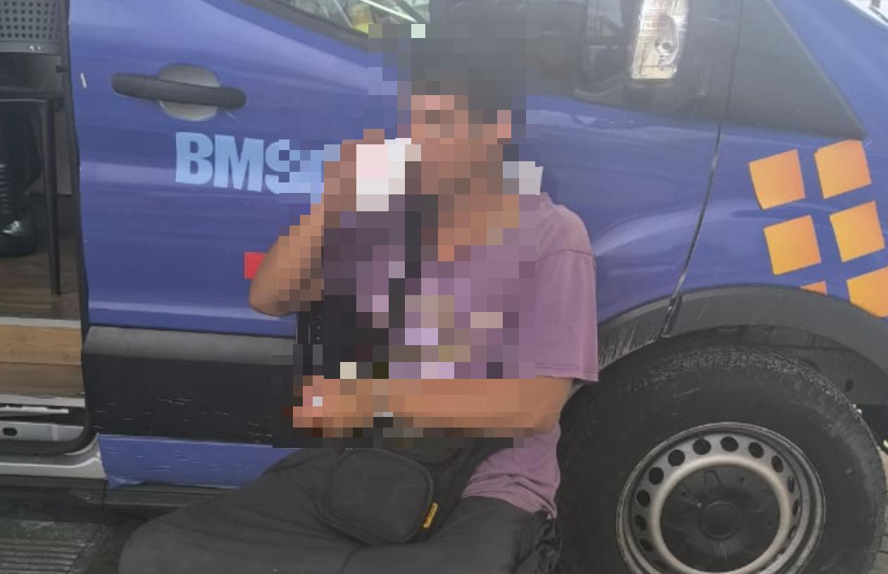 A imagem mostra um homem colombiano que fugiu de fazendo em Maracanaú, com a identidade preservada e fisionomia borrada, vestindo blusa roxa e calça preta, com uma bolsa preta, se alimentando próximo a um carro azul.