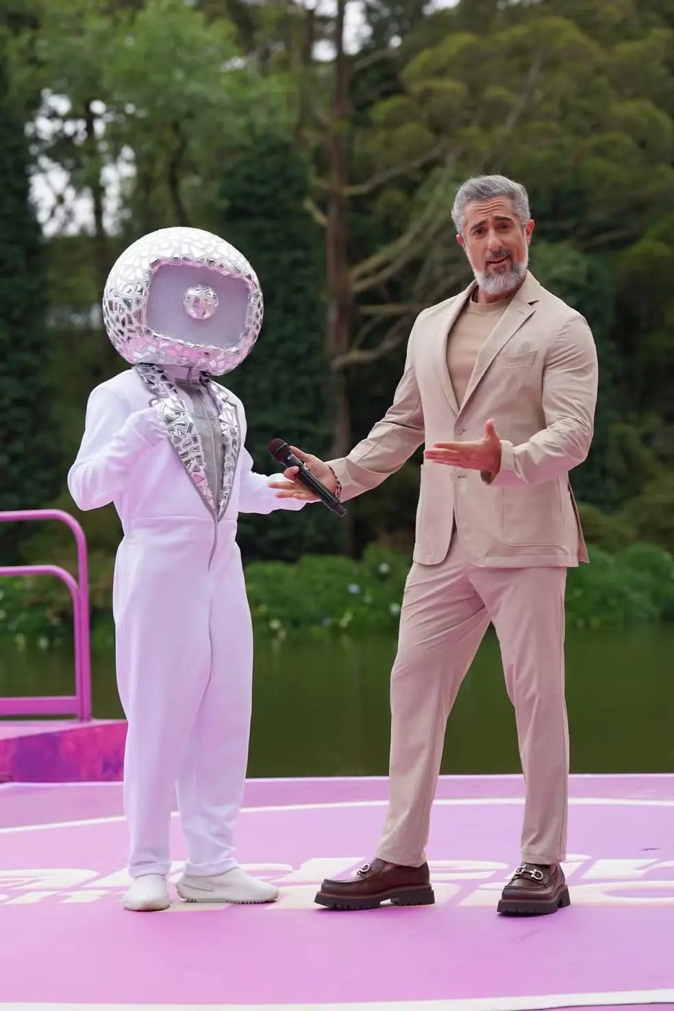 A imagem mostra o apresentador Marcos Mion e outra pessoa, que utiliza uma cabeça representativa da TV Globo.