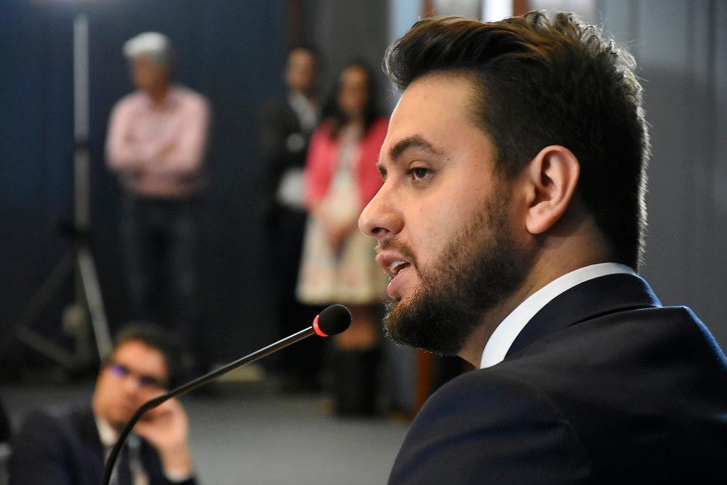 Homem branco de cabelo, barba e bigode castanho escuro, usando um termo e olhando de perfil para o lado esquerdo. Um microfone está perto dele.
