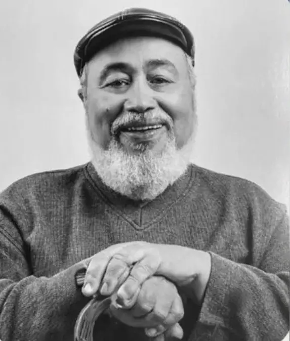 Imagem em preto e branco de um rosto de homem negro, idoso, com barba e usando chapéu.