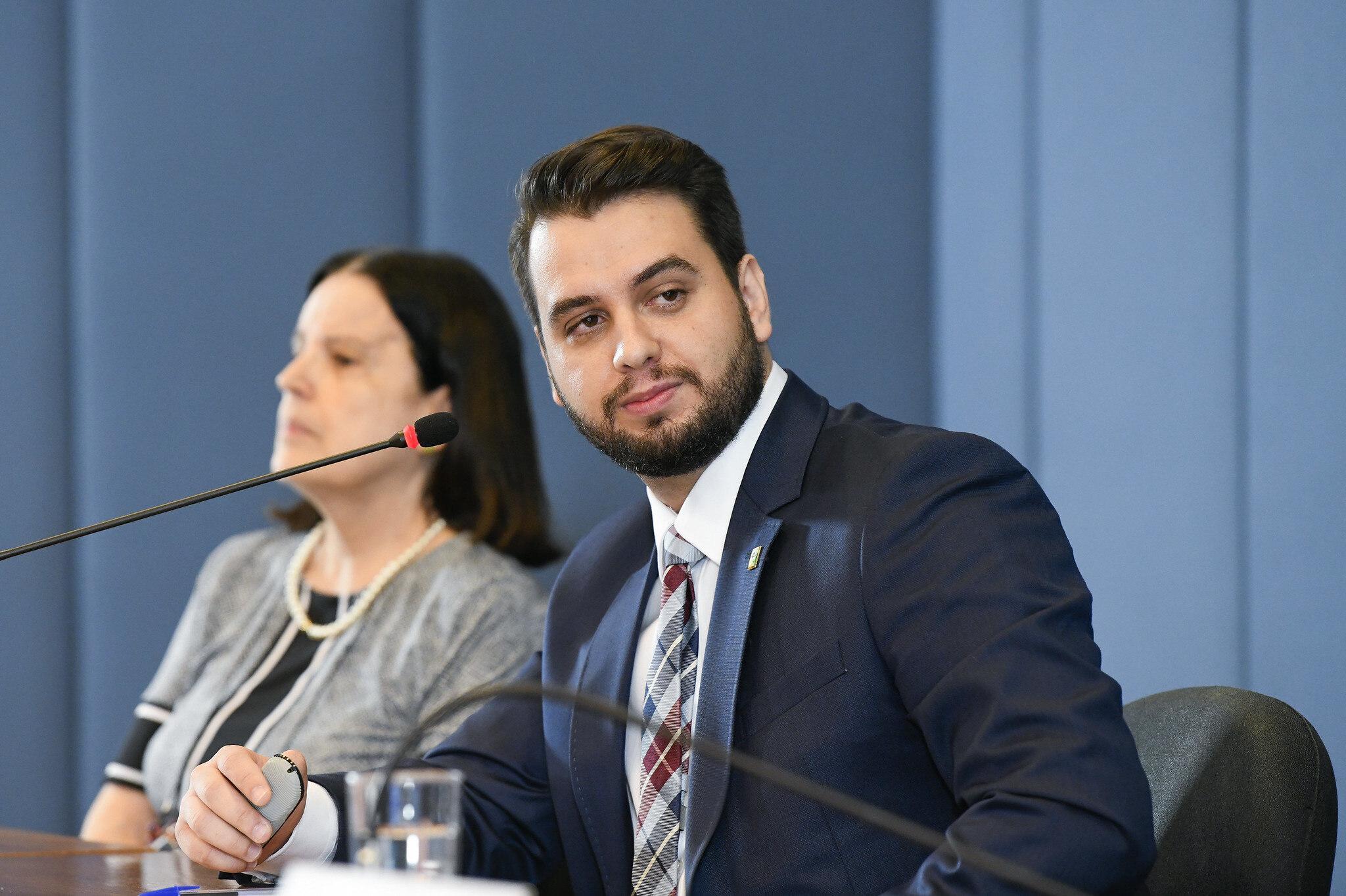 Filipe Martins usa terno e gravata, sentado à mesa com um microfone, participa de palestra; ao fundo, uma mulher também sentada diante de um microfone, em ambiente institucional. Imagem usada para informar quem é ele.