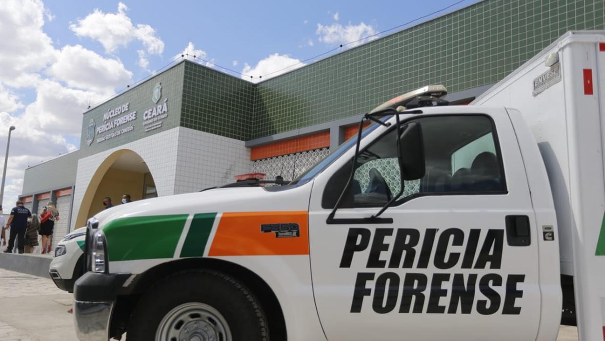 Viatura branca da Perícia Forense com faixas verde e laranja estacionada em frente a um prédio público identificado como Núcleo de Perícia Forense do Ceará. Ao fundo, pessoas circulam pela entrada do edifício, que tem fachada revestida de azulejos verdes, sob céu parcialmente nublado.