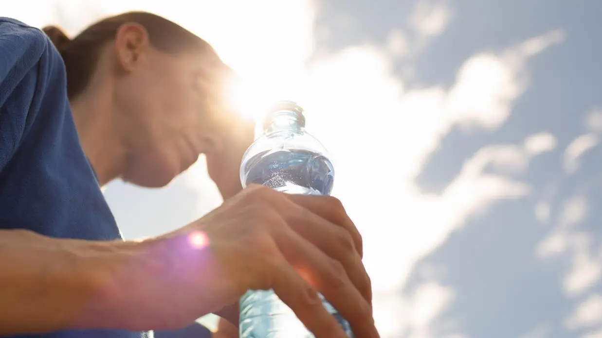 A imagem mostra uma mulher, vestida de blusa azul, aparentemente com calor, segurando uma garrafa de água, em um dia bastante ensolarado.