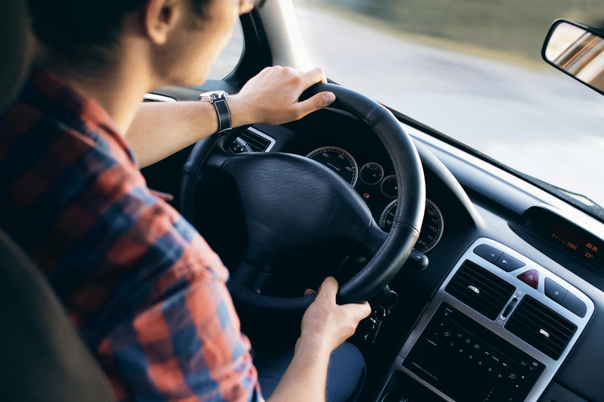 Foto dentro de carro de homem de camisa xadrez vermelha e azul com mãos no volante.