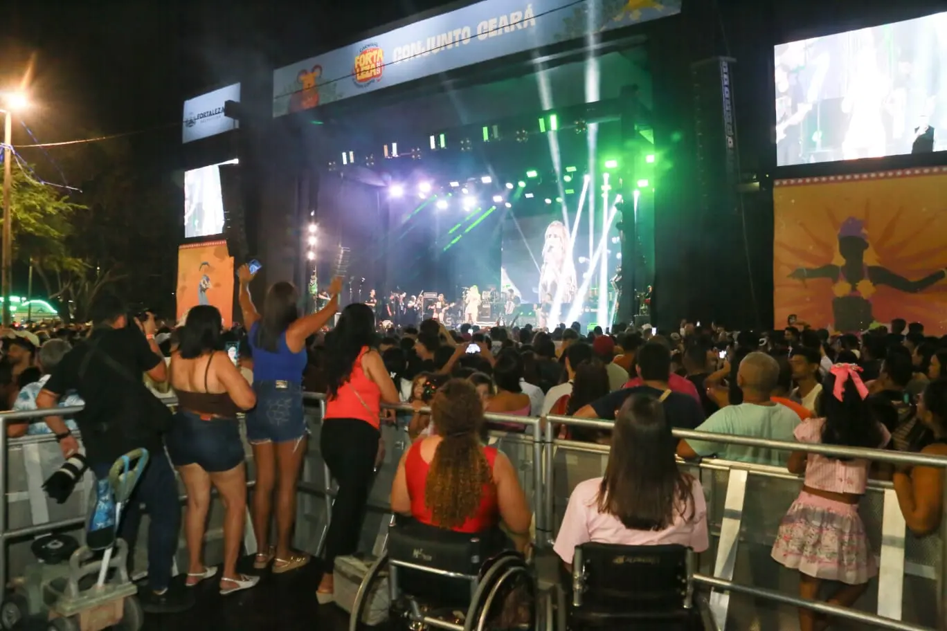 Foto de festa pública no bairro Conjunto Ceará, em Fortaleza. Centenas de pessoas estão próximas a um palco com luzes e uma cantora se apresentando. Em primeiro plano, há uma área com grades para pessoas com deficiência em cadeiras de rodas assistirem à apresentação.