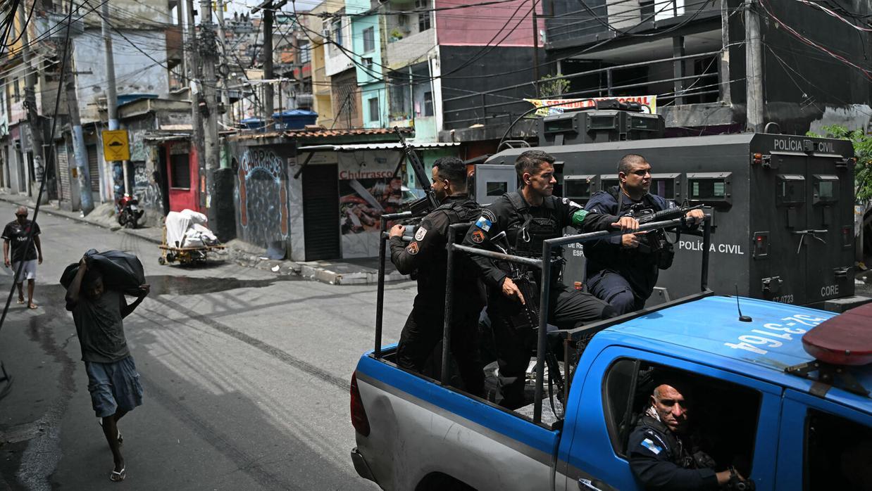 A foto mostra policiais militares em cima e dentro de um carro da Polícia do Rio de Janeiro, fortemente armados, passando por uma comunidade, enquanto pessoas trafegam pela via.