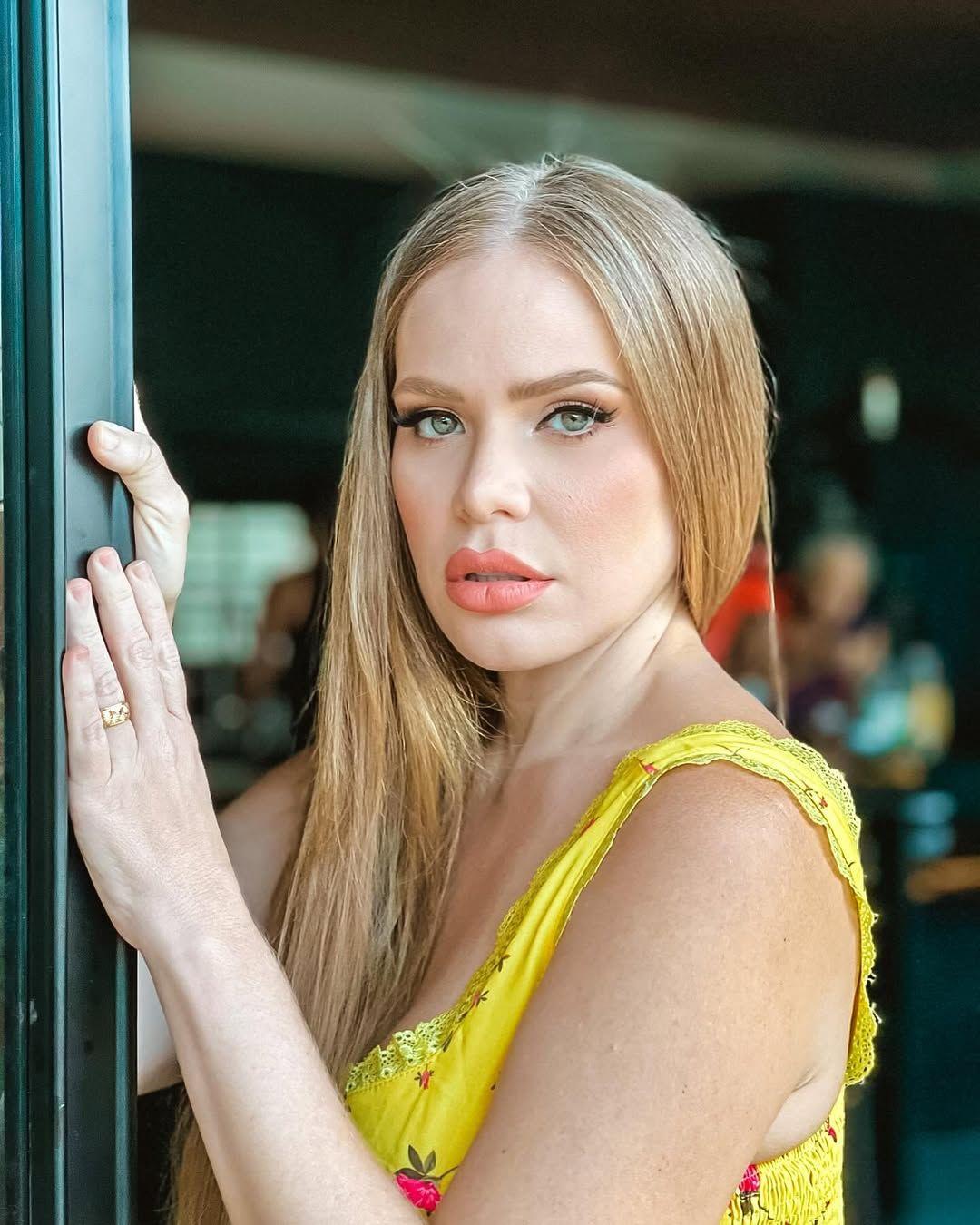 Paulinha Leite com camiseta amarela na entrada de uma porta segurando a porta com uma das mãos.