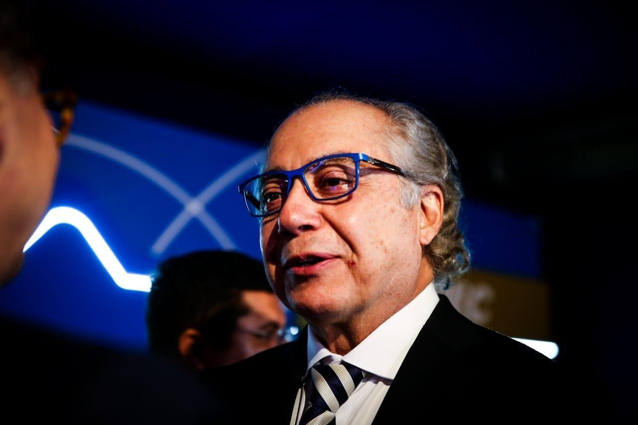 Esta imagem mostra o plano médio de Mário Araripe, um homem de pele clara e meia-idade, com cabelos grisalhos e ondulados que chegam até a altura do pescoço, usando óculos de armação azul vibrante que se destacam em seu rosto. Ele está levemente voltado para a esquerda, com uma expressão serena e a boca entreaberta como se estivesse em meio a uma fala, vestindo um terno preto clássico, camisa branca e uma gravata de listras diagonais em tons de azul e branco. O fundo é composto por um painel azul com detalhes de linhas curvas brancas iluminadas e uma parte dourada à direita, criando um ambiente formal e bem iluminado que realça os detalhes de seu rosto e o brilho em seus olhos.