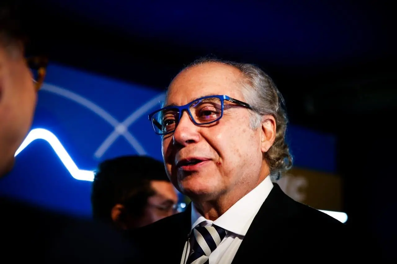 Esta imagem mostra o plano médio de Mário Araripe, um homem de pele clara e meia-idade, com cabelos grisalhos e ondulados que chegam até a altura do pescoço, usando óculos de armação azul vibrante que se destacam em seu rosto. Ele está levemente voltado para a esquerda, com uma expressão serena e a boca entreaberta como se estivesse em meio a uma fala, vestindo um terno preto clássico, camisa branca e uma gravata de listras diagonais em tons de azul e branco. O fundo é composto por um painel azul com detalhes de linhas curvas brancas iluminadas e uma parte dourada à direita, criando um ambiente formal e bem iluminado que realça os detalhes de seu rosto e o brilho em seus olhos.