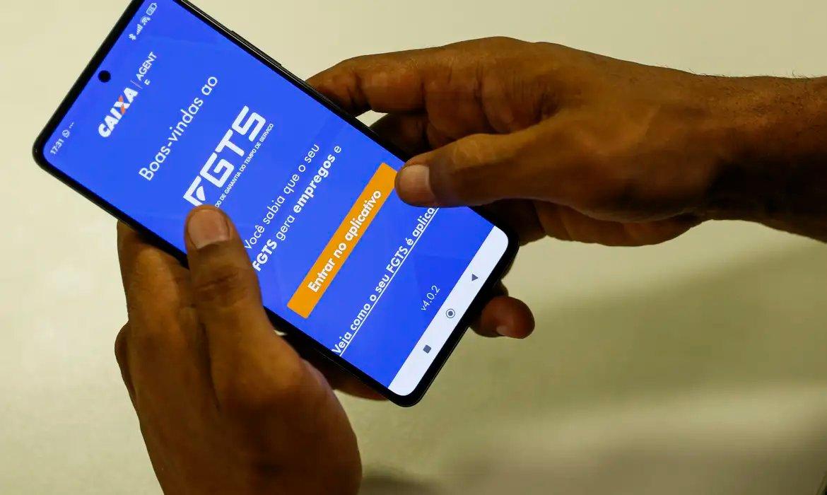 Close-up de mãos segurando um smartphone com o aplicativo FGTS da Caixa aberto na tela de boas-vindas, exibindo a opção Entrar no aplicativo.