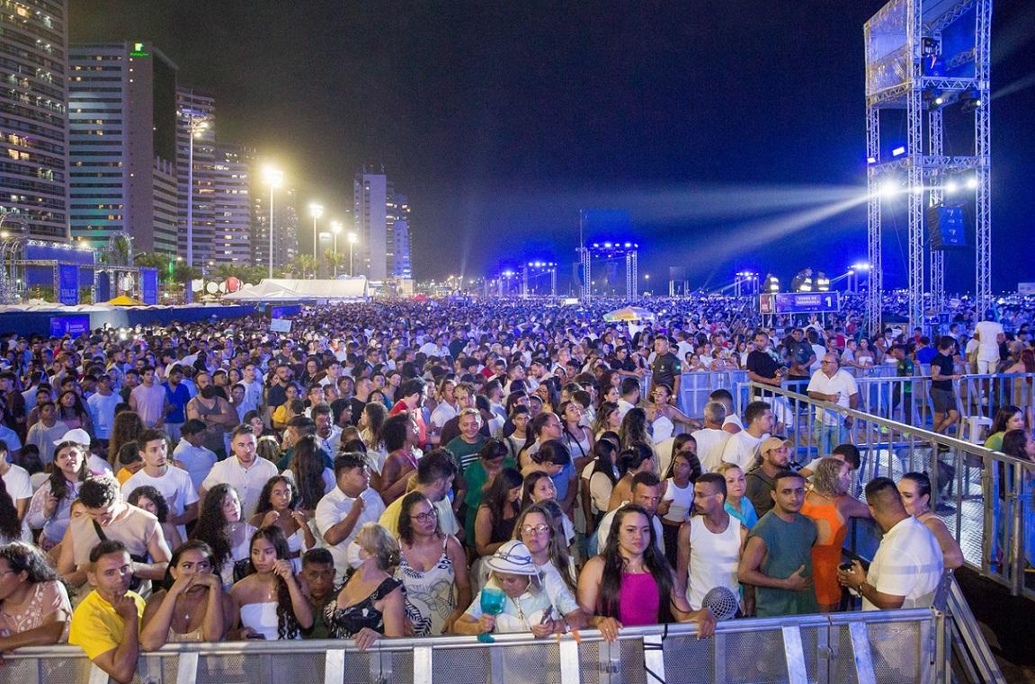 Vasta multidão reunida em um show noturno ao ar livre no aterro da praia de Iracema, com um palco iluminado por intensas luzes azuis e estruturas metálicas visíveis ao fundo, ladeado por edifícios urbanos.
