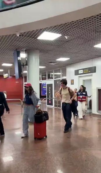 Bruna Marquezine e Shawn Mendes caminhando em um terminal de aeroporto, com malas e mochilas, em um ambiente interno iluminado.