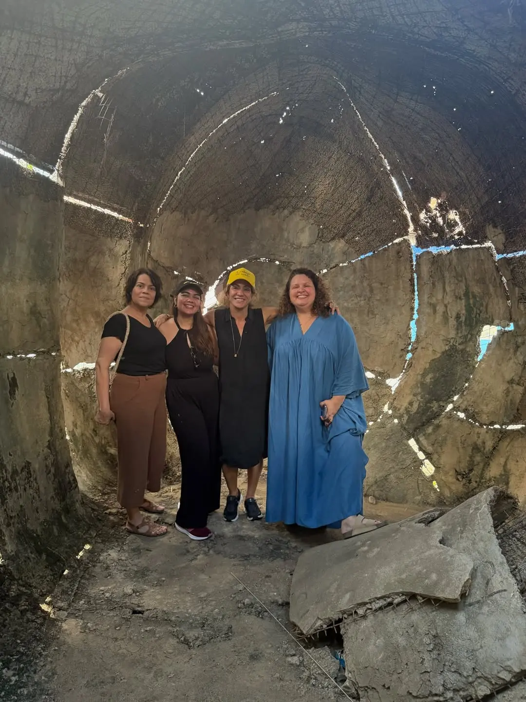 Quatro mulheres em pé com uma estrutura de pedra ao fundo.