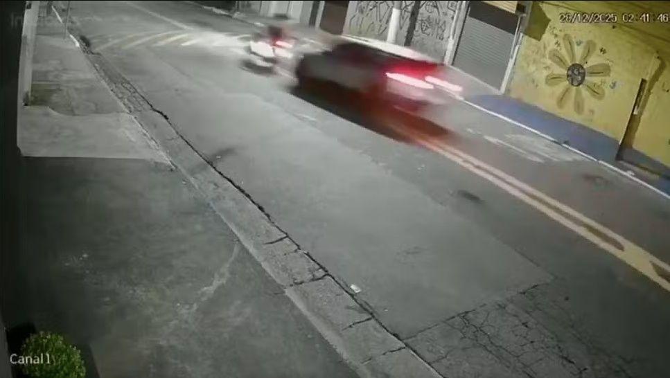 Câmera de segurança noturna registrando um carro em alta velocidade com luzes traseiras vermelhas borradas e uma motocicleta à frente em uma rua urbana escura.