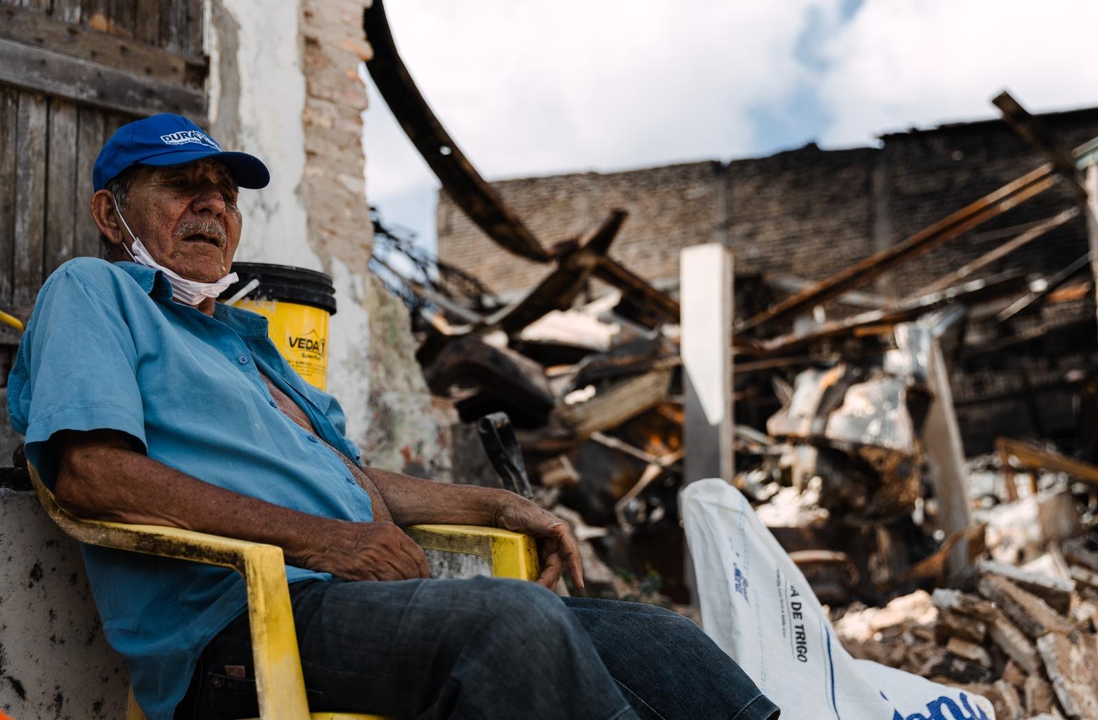 Chico Alves, um homem idoso de boné azul e camisa azul, com uma máscara no pescoço, sentado em uma cadeira amarela, com ruínas de uma construção destruída e céu nublado ao fundo.