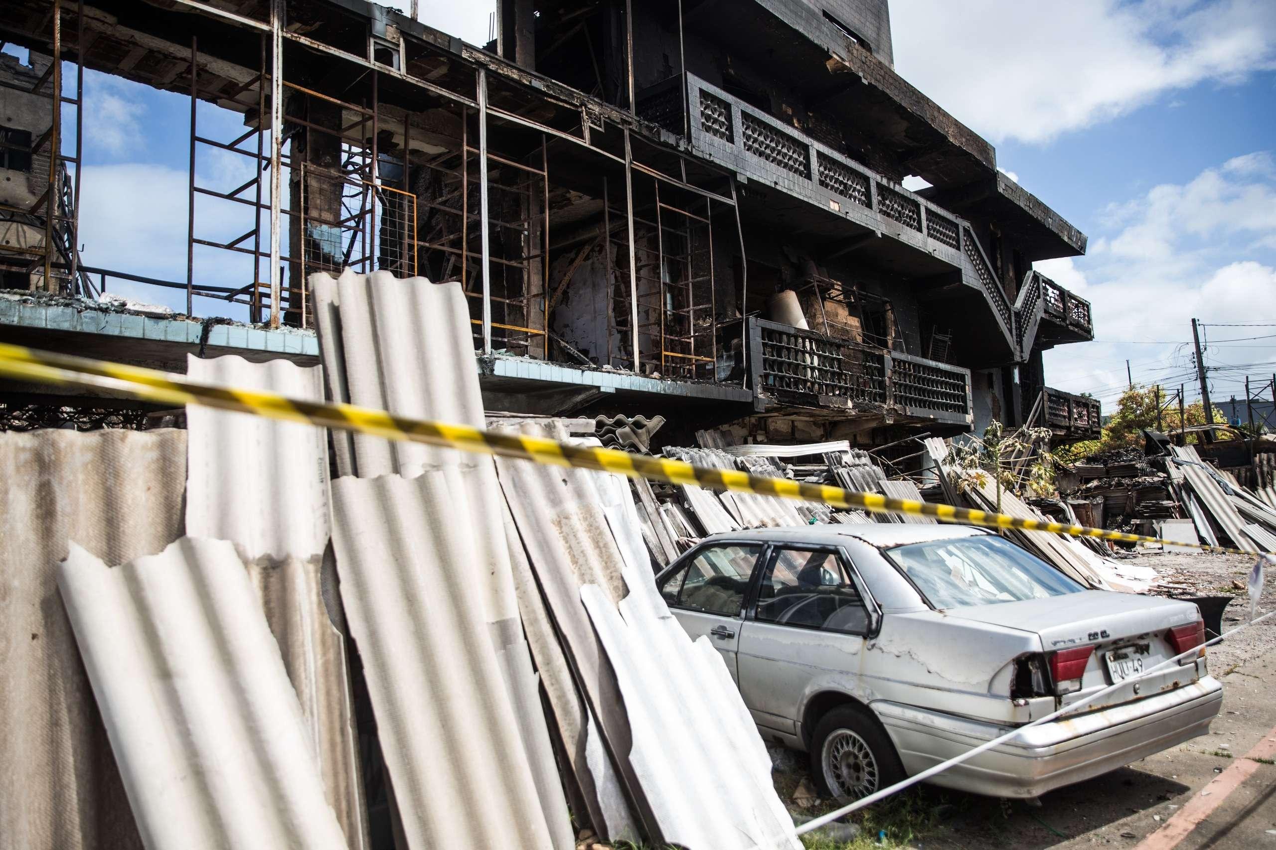 Vista da fachada da sucata Chico Alves, severamente danificada por incêndio, com a estrutura carbonizada e janelas quebradas. Em primeiro plano, há uma fita de isolamento amarela e preta, telhas de fibrocimento quebradas e um carro prateado amassado.