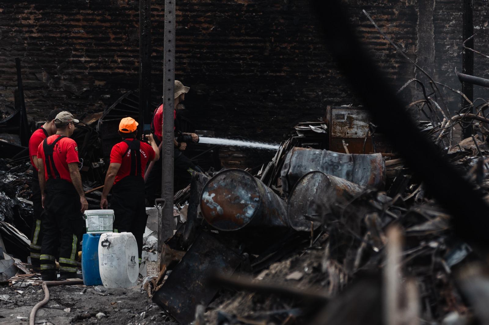 Bombeiros em uniformes vermelhos combatem rescaldo de incêndio, pulverizando água sobre destroços carbonizados e barris metálicos na sucata Chico Alves, em Fortaleza.