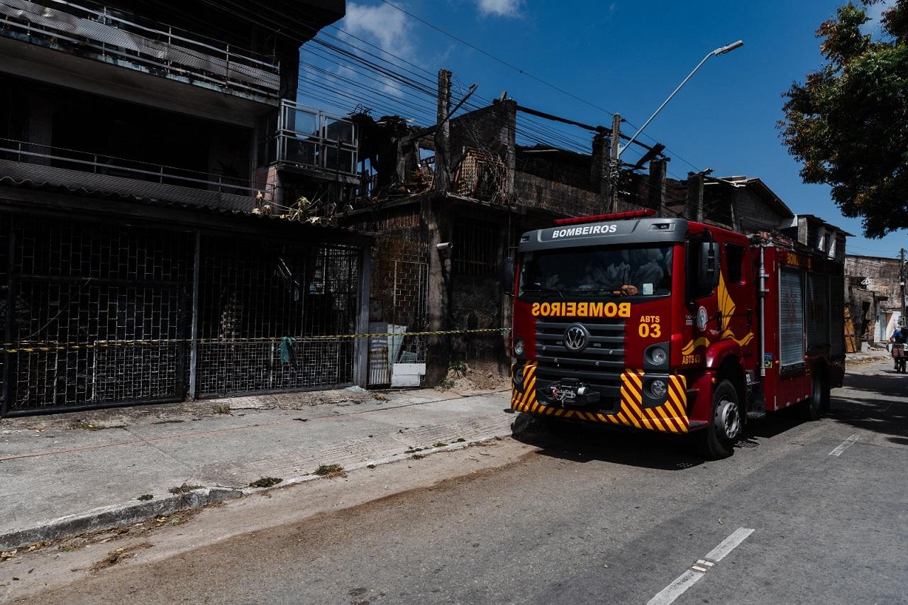 Caminhão vermelho do Corpo de Bombeiros do Ceará estacionado em frente a edifícios visivelmente danificados e carbonizados por um incêndio, com fita de isolamento amarela, no bairro Jacarecanga.