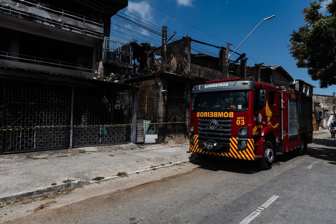 Caminhão vermelho do Corpo de Bombeiros do Ceará estacionado em frente a edifícios visivelmente danificados e carbonizados por um incêndio, com fita de isolamento amarela, no bairro Jacarecanga.