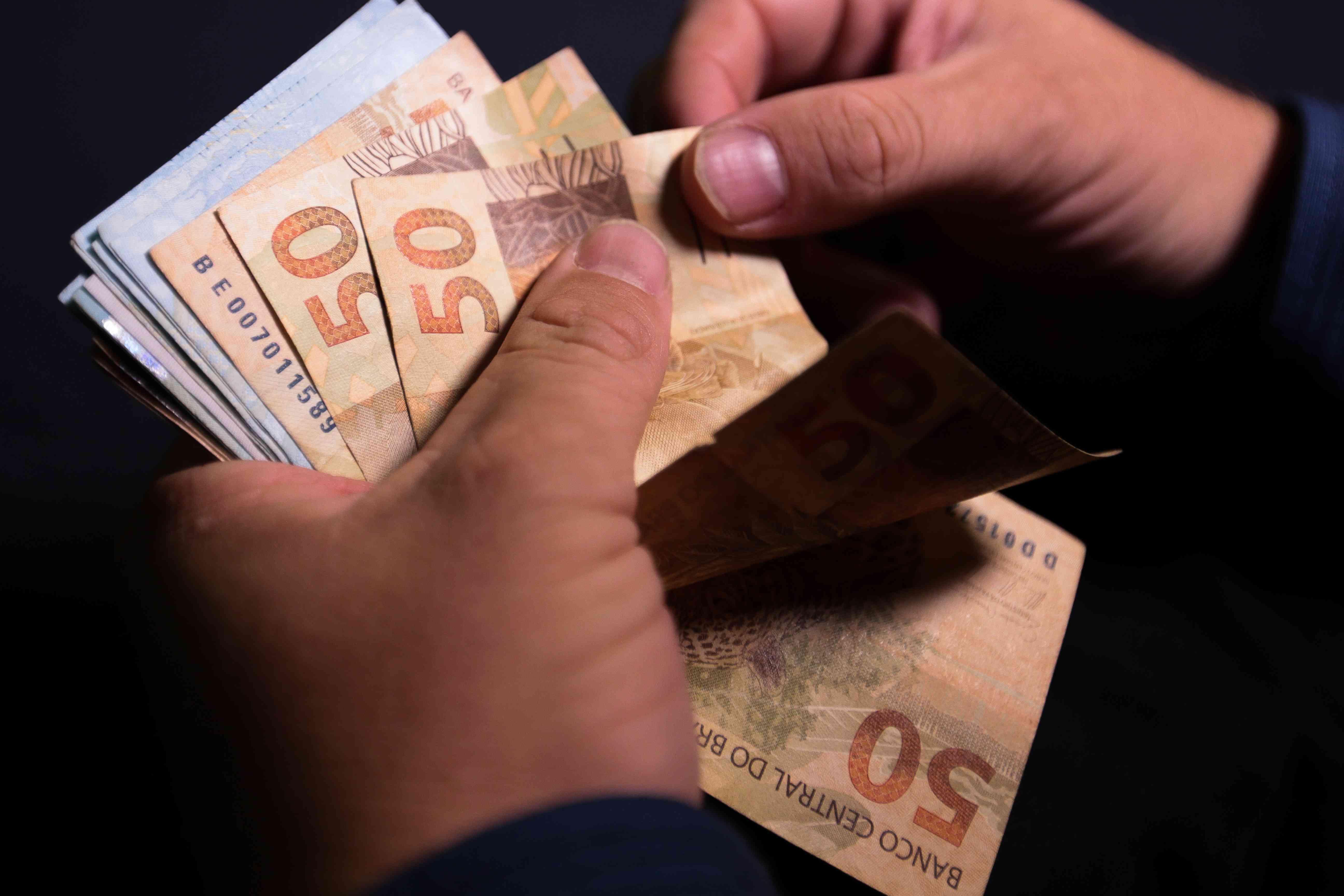 Mãos contando notas de dinheiro em reais, incluindo cédulas de 50, representando finanças pessoais e gestão de dinheiro no Brasil.
