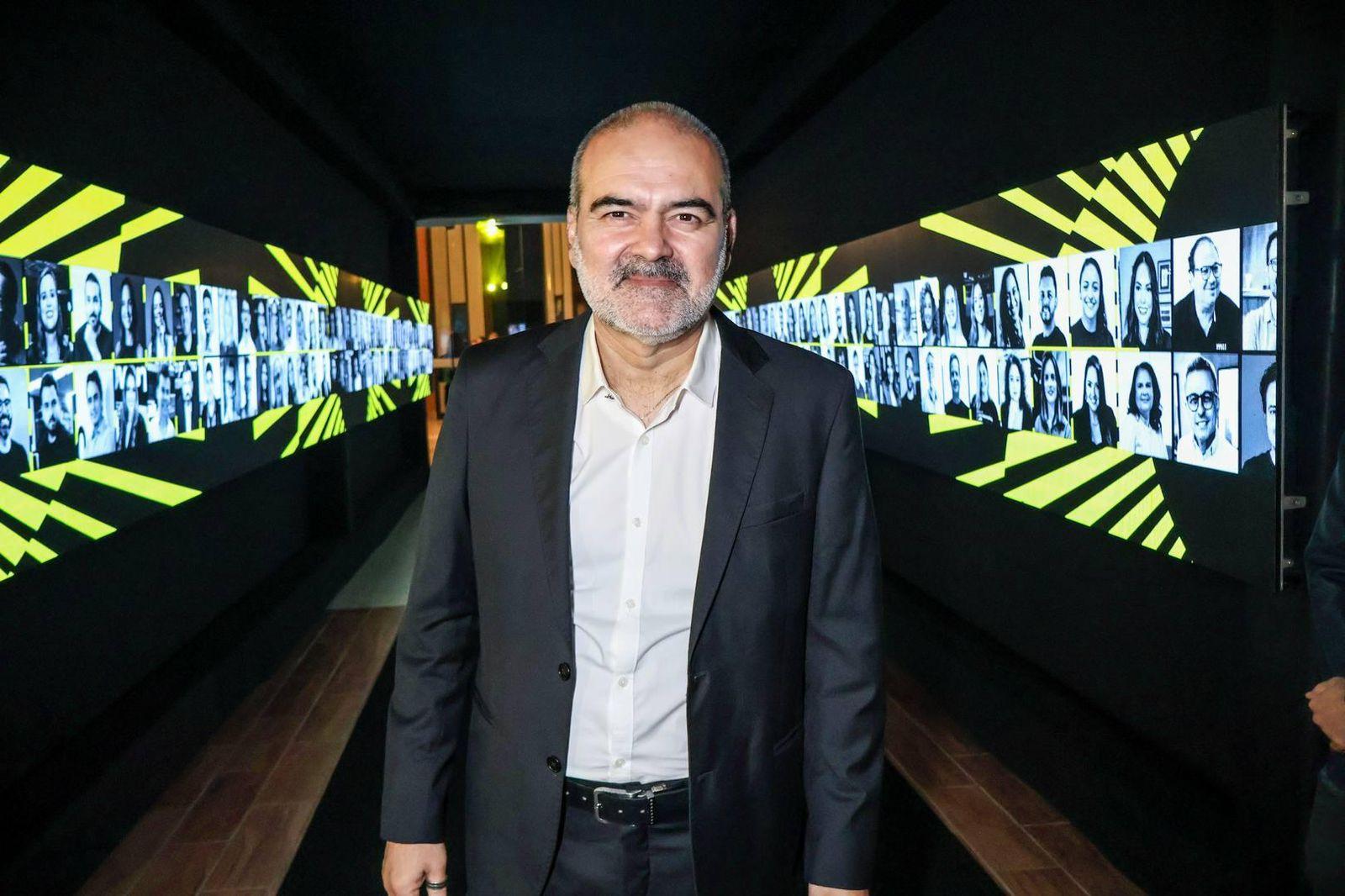 Homem de meia-idade, usando terno escuro e camisa branca, posa no centro de um corredor escuro iluminado por telas laterais. Nas paredes à esquerda e à direita, há dezenas de retratos em preto e branco de diferentes pessoas, dispostos em sequência, intercalados por elementos gráficos em amarelo. Ele olha diretamente para a câmera, com expressão serena, em um ambiente que remete a evento corporativo ou exposição audiovisual.