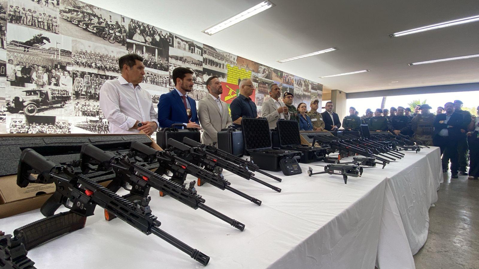 Mesa coberta com tecido branco exibindo diversos fuzis pretos alinhados, caixas de transporte rígidas abertas e fechadas, além de um drone. Ao fundo, há um grupo de pessoas em pé, algumas usando trajes formais e outras uniformes, diante de uma parede decorada com fotos históricas em preto e branco e um painel com texto em destaque.