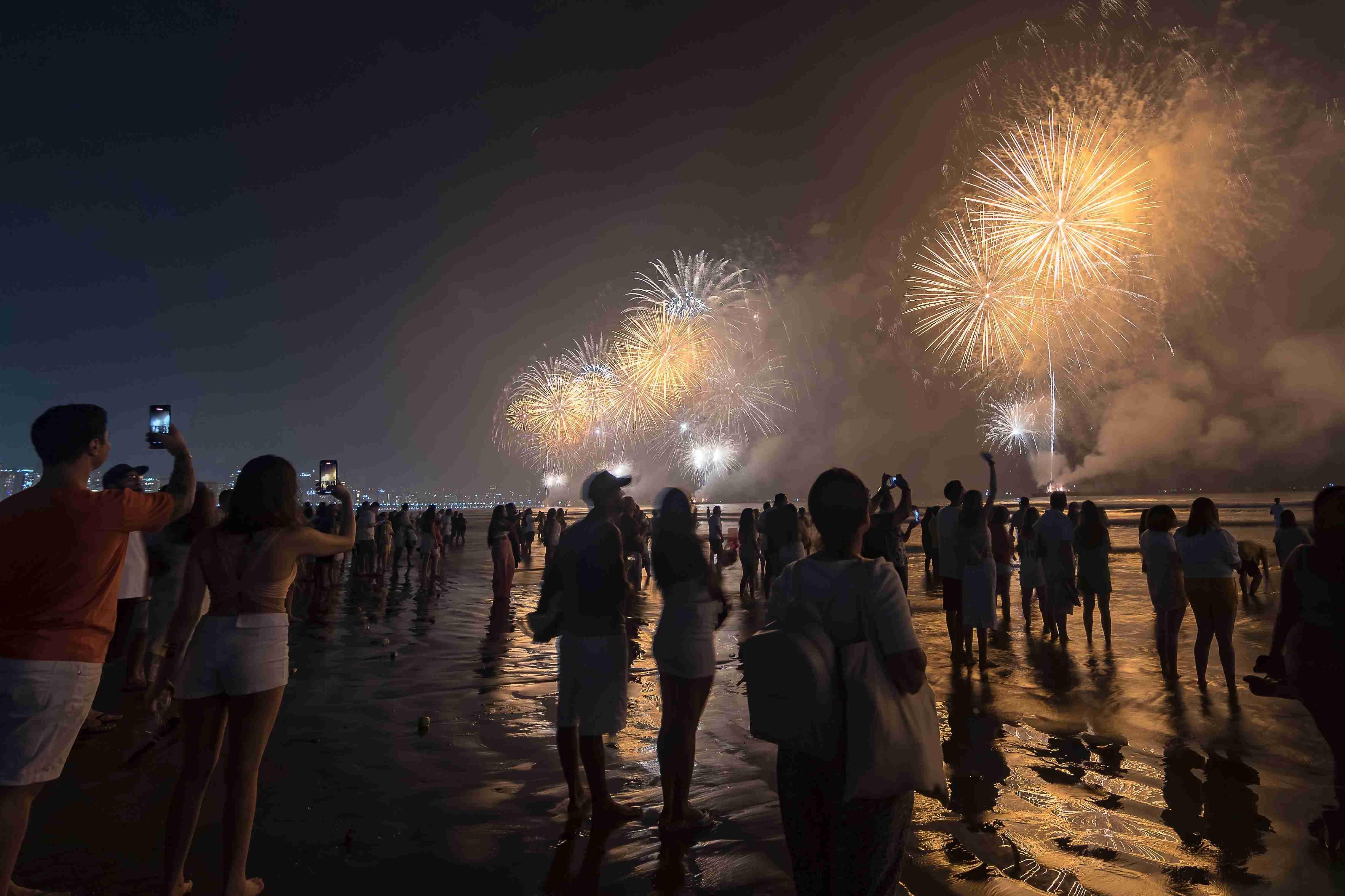 Multidão em uma praia noturna observando e filmando um grandioso espetáculo de fogos de artifício dourados, durante o Ano Novo, com reflexos na areia molhada.