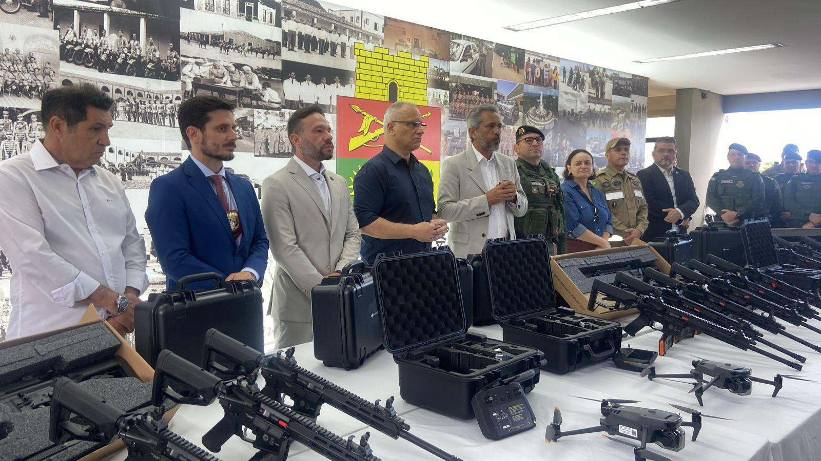 A imagem mostra homens e uma mulher, autoridades cearenses, atrás de uma mesa, onde são expostas armas de fogo de grosso calibre e drones.