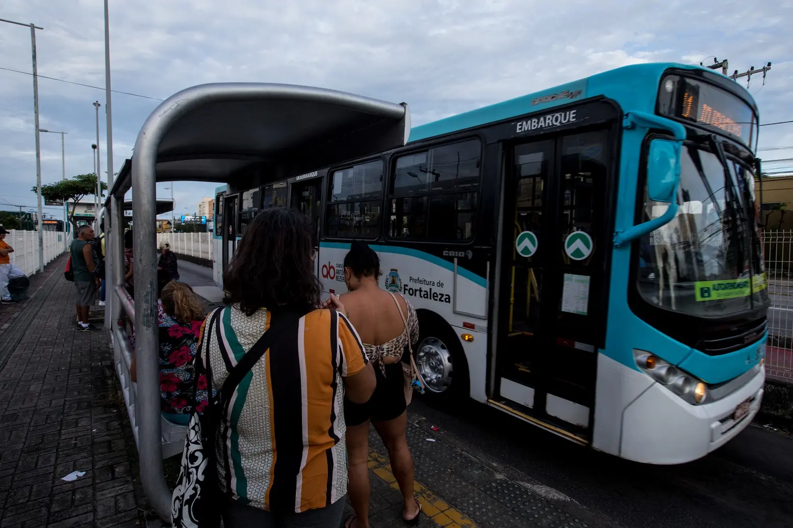Mulheres aguardando em um ponto de ônibus urbano em Fortaleza, com um ônibus azul e branco parado para embarque.