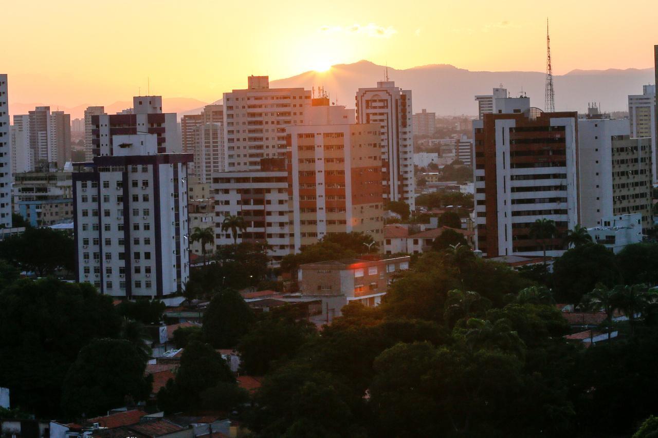 Vista panorâmica de Fortaleza ao pôr do sol, com numerosos prédios residenciais e comerciais, árvores escuras em primeiro plano e montanhas ao fundo, onde o sol se põe, emitindo raios dourados.