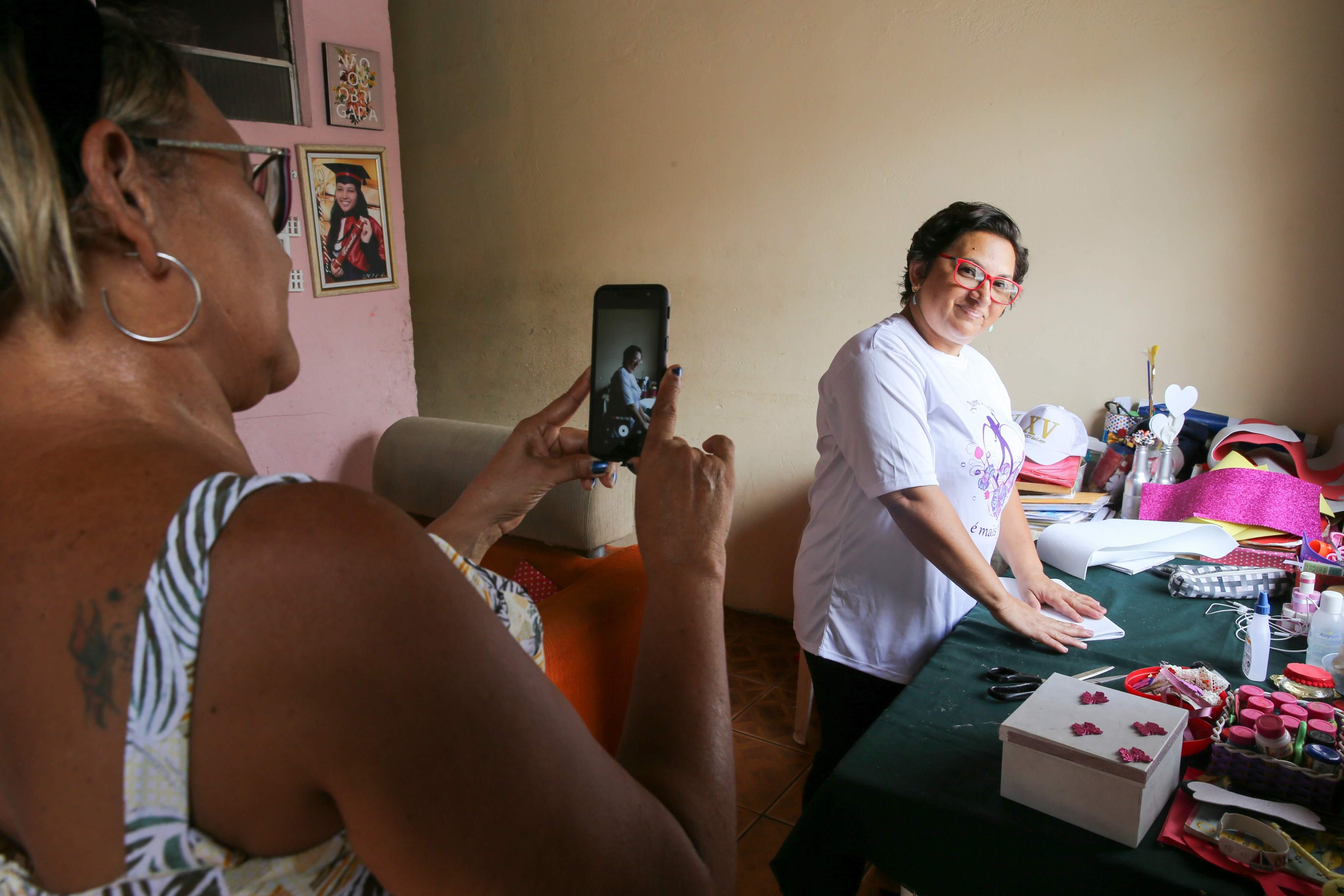 Mulher de óculos vermelhos e camiseta branca sorri enquanto é filmada por um celular em uma mesa repleta de materiais de artesanato. Em primeiro plano, aparecem as mãos e as costas de outra mulher segurando o aparelho para realizar a gravação.