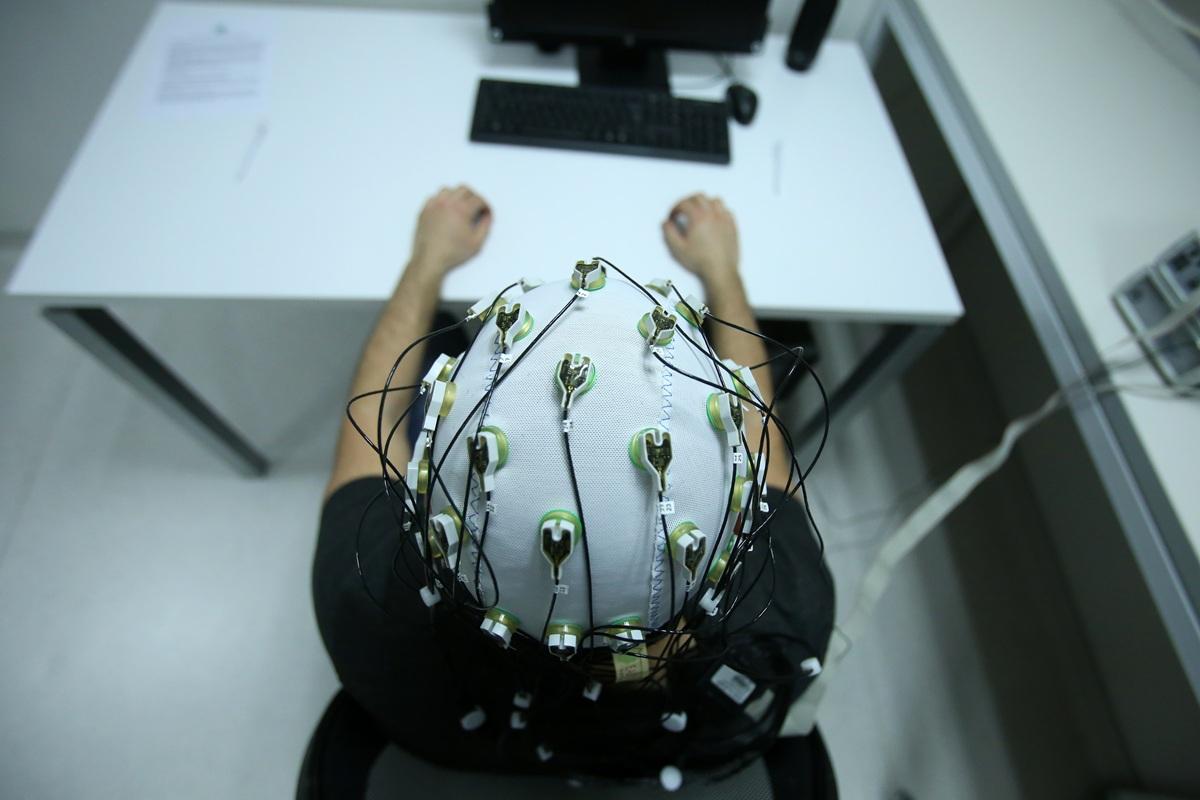 Pessoa sentada à mesa, vista de cima, usando uma touca com eletrodos e fios para monitoramento da atividade cerebral (EEG), com as mãos apoiadas à frente de um computador.