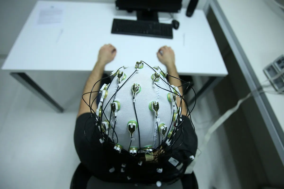 Pessoa sentada à mesa, vista de cima, usando uma touca com eletrodos e fios para monitoramento da atividade cerebral (EEG), com as mãos apoiadas à frente de um computador.