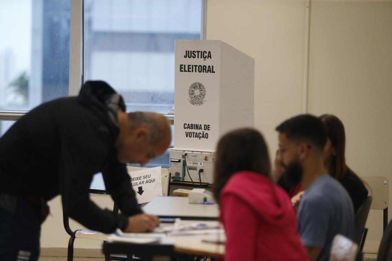 Foto mostra atendentes e eleitor antes de entrar na cabine para votar na urna eletrônica do Tribunal Superior Eleitoral (TSE).