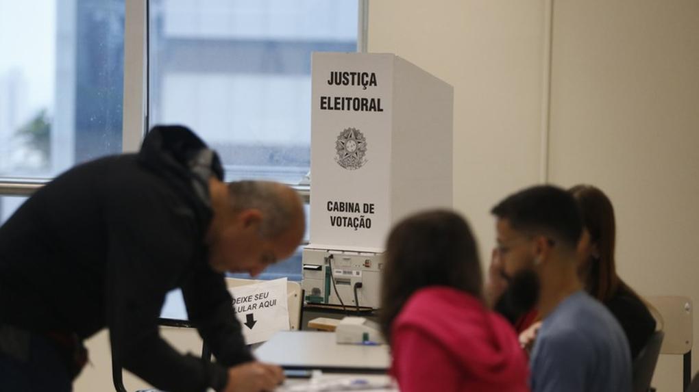 Foto mostra atendentes e eleitor antes de entrar na cabine para votar na urna eletrônica do Tribunal Superior Eleitoral (TSE).