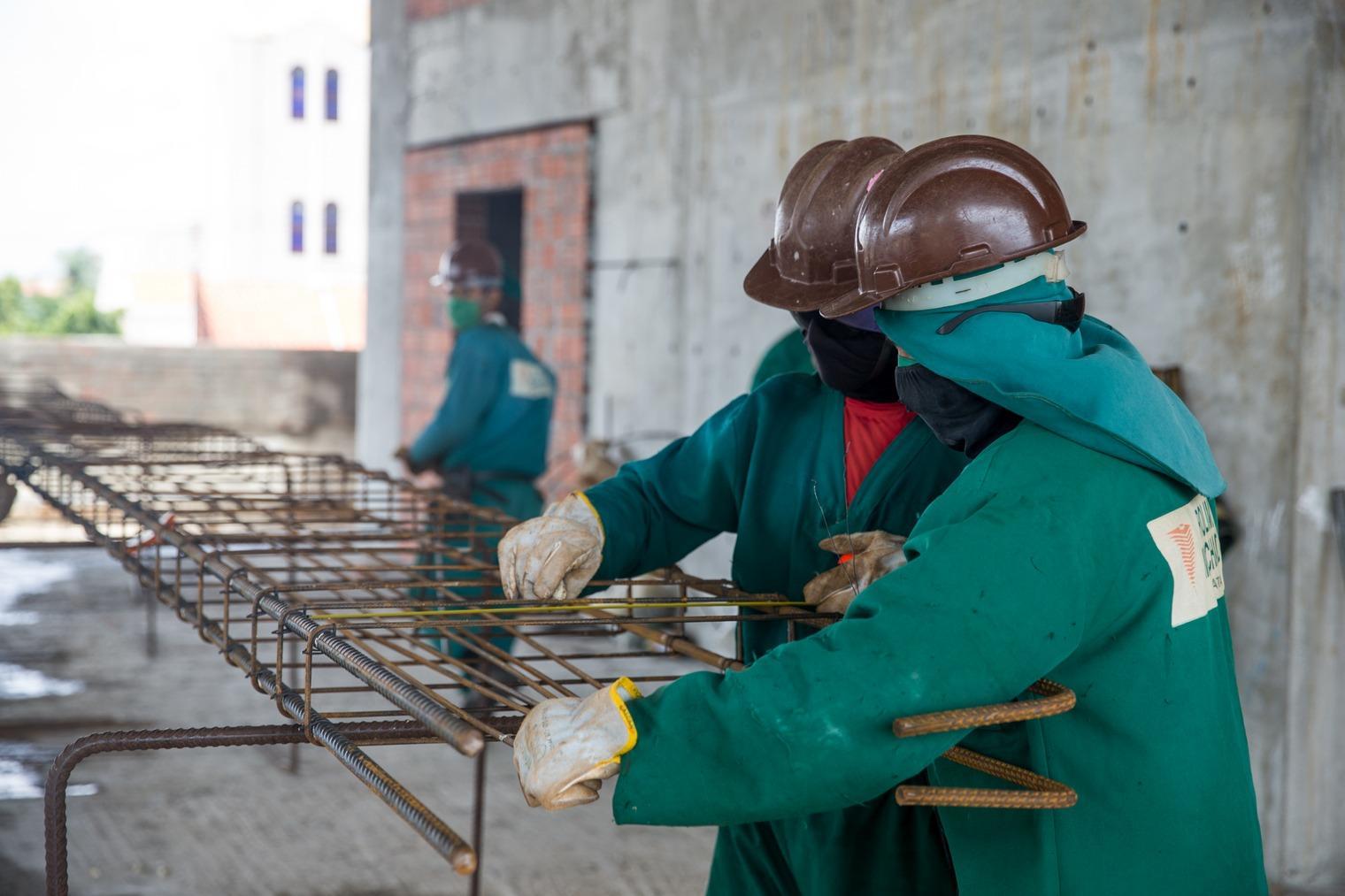 Trabalhadores da construção civil usam capacetes e equipamentos de proteção enquanto montam armações de ferro em obra de edifício.