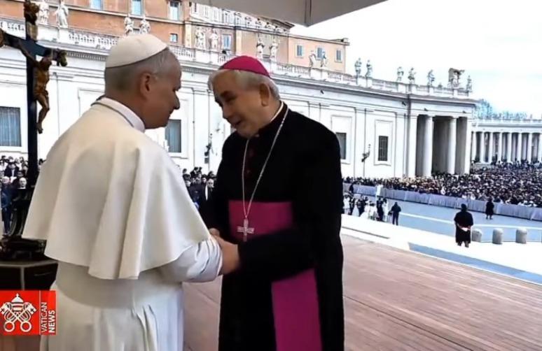 Dois líderes religiosos se cumprimentam em um palco ao ar livre, diante de uma multidão reunida na Praça de São Pedro, no Vaticano.