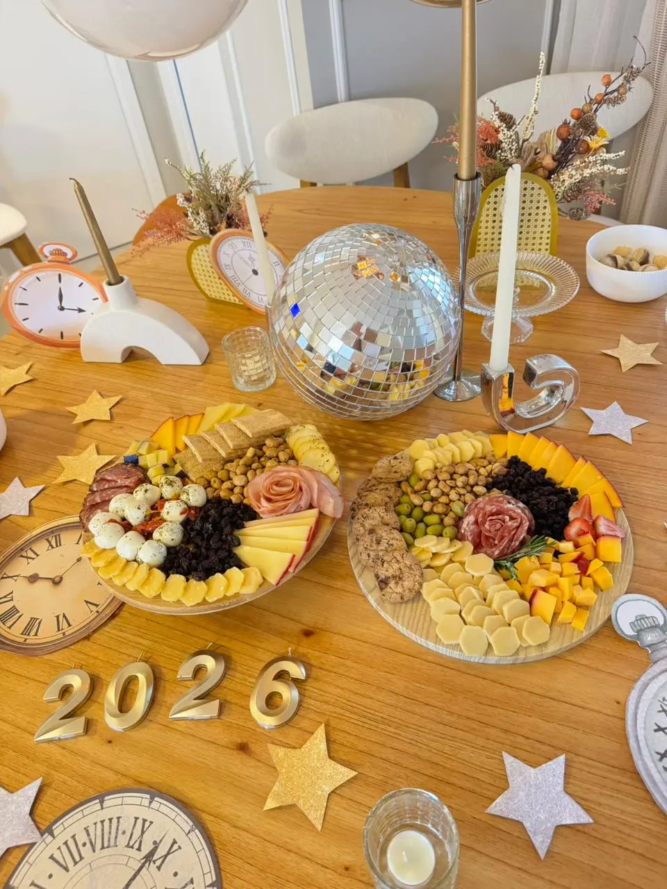 Uma mesa de madeira decorada para o Réveillon 2026 apresenta duas tábuas de frios fartas com queijos, embutidos em formato de rosa e petiscos ao redor de um globo espelhado central. Elementos temáticos como relógios, estrelas douradas e velas completam a composição festiva e iluminada sob um tom rústico e sofisticado.
