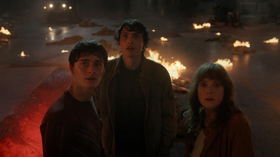 Três pessoas em pé em um espaço escuro, cercadas por chamas espalhadas pelo chão, com um veículo iluminado ao fundo.